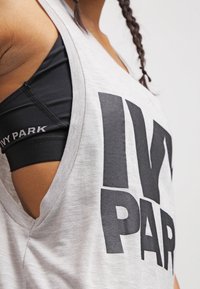 Szary top na ramiączkach z dużym czarnym napisem "IVY PARK", z bezrękawowym fasonem i otwartym bokiem dla lepszej wentylacji. Czar bra sportowy widoczny pod spodem.