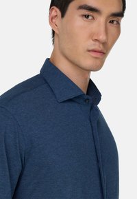 Blauwe button-up blouse met een gestructureerde kraag, textuurstof en subtiel heatherpatroon. Heeft een slim fit en een enkele zwarte knoop.