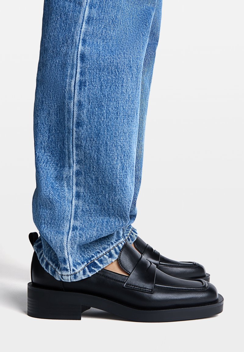 Mocassins en cuir noir avec un petit talon carré, présentant un design à bande croisée. Portés avec un jean bleu clair légèrement délavé.