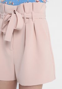 Svetle růžové šortky s vysokým pasem vyrobené z texturované tkaniny, s páskem na mašličku, záhyby a bočními kapsami pro funkčnost.