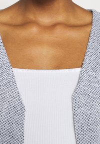 Zbliżenie na szyję i górną część klatki piersiowej osoby noszącej białą prążkowaną bluzkę i szary teksturowany kardigan rozpinany.