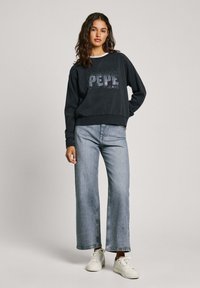 Sudadera negra con texto plateado "PEPE JEANS", cuello y puños acanalados, combinada con jeans de pierna ancha en azul claro y zapatillas blancas.