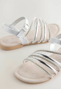 Zilveren sandalen met bandjes en een glanzende metalen afwerking, voorzien van rhinestone-accenten op een zachte, suede-achtige beige voetbed. Platte zool.