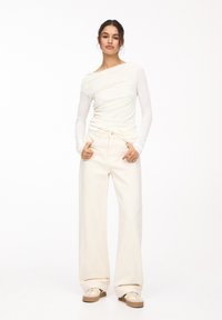 Haut blanc à manches longues avec un design drapé, associé à un pantalon crème à jambes larges et des baskets beiges. Textures lisses et style minimaliste.