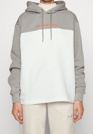 Osoba nosząca szaro-białą bluzę z kapturem w blokowe kolory z napisem "Calvin Klein Jeans" w kolorze pomarańczowym na piersi oraz beżowe spodnie z haftowanym logo.