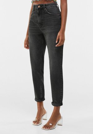 Hoge taille zwarte denim jeans met een taps toelopende pasvorm, opgerolde boorden en contrasterende stiksels. Gedragen met doorzichtige hoge hakken.