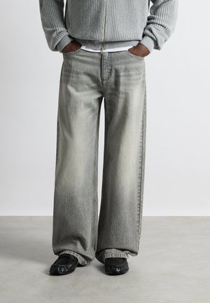 Personne portant un jean gris ample et délavé, des chaussures noires et un pull gris zippé avec les mains dans les poches, debout sur un sol gris.