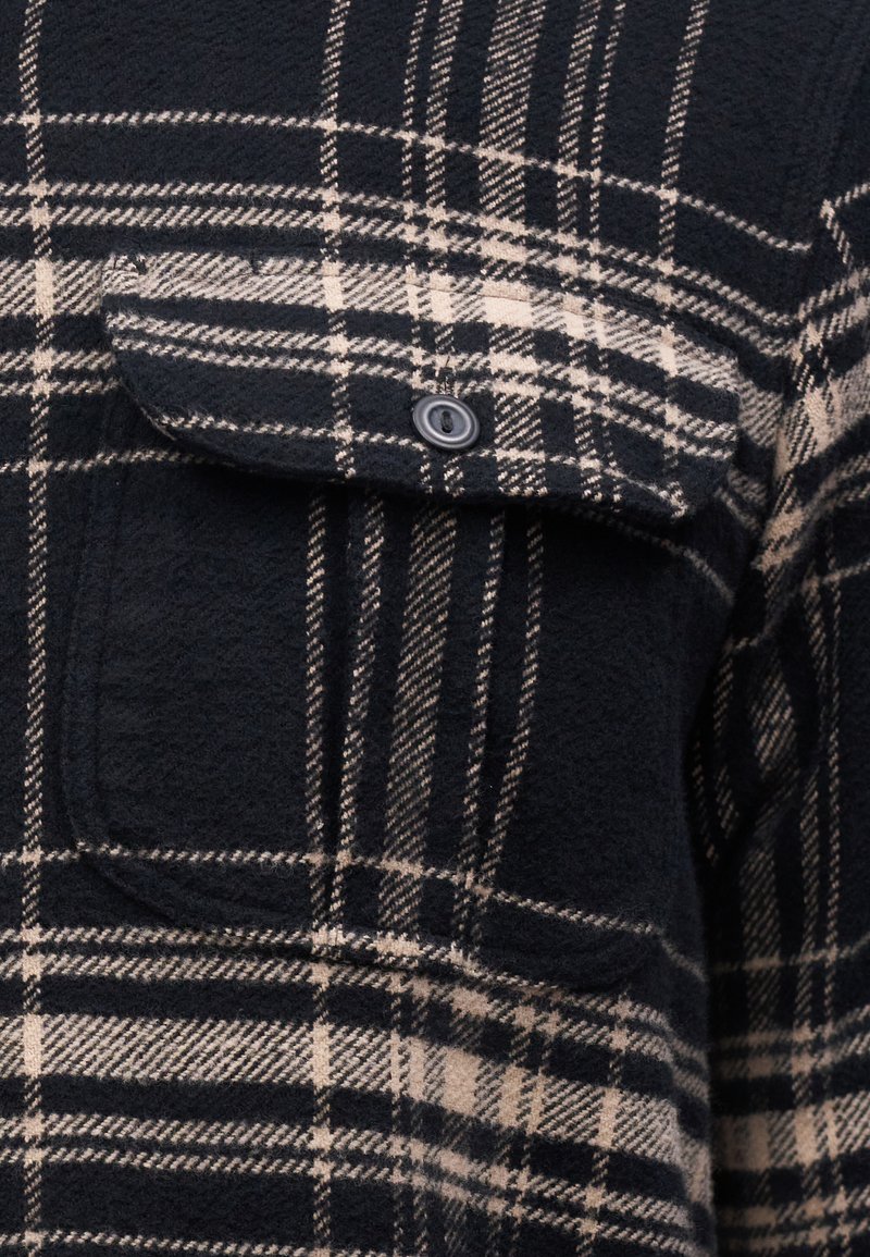 Camisa de franela a cuadros en negro y beige con un bolsillo en el pecho, que presenta una tela texturizada y un cierre de botón en el bolsillo.