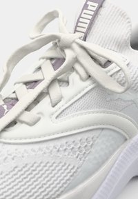 Chaussure de sport blanche avec tige en mesh texturé, superpositions lisses et accents violets. Elle dispose d'une semelle rembourrée et de lacets plats.