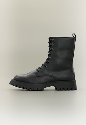 Bota de combate negra de cuero con cordones, suela gruesa con estrías y ojales de metal, vista de lado sobre un fondo liso.
