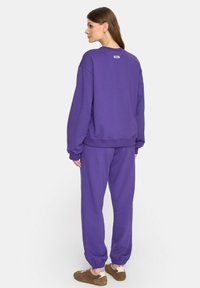 Kobieta stojąca bokiem, ubrana w fioletową bluzę i pasujące dresy, w brązowych trampkach na białym tle.