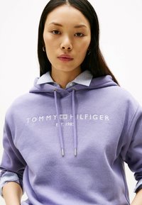 Paarse hoodie met wit "TOMMY HILFIGER" logo en "EST. 1985" tekst. Bevat een trekkoord capuchon en een gelaagde look met een blauw-wit gestreept shirt eronder.
