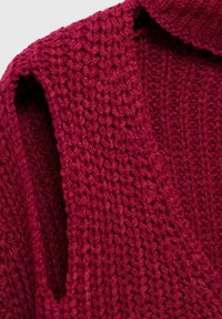 Pletený odev v hlbokej červenej farbe s hrubou textúrou. Má široký golier a bezrukávový dizajn, ktorý ukazuje voľné, otvorené stehy.