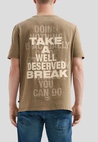 Une personne portant un t-shirt marron avec un texte en gras "FAITES UNE PAUSE BIEN MÉRITÉE" dans le dos, associé à un jean bleu.