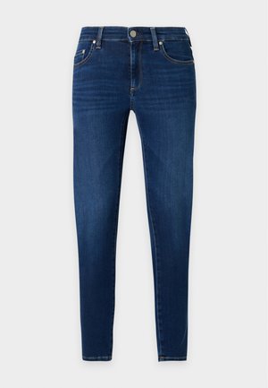 Donna con capelli neri che indossa un top grigio senza maniche, jeans skinny blu scuro e ballerine nere, in piedi con una mano in tasca.