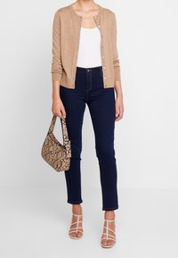 Béžový cardigan, bílý top na ramínka, tmavě modré džíny a sandály na pásku. Doplňky zahrnují hnědou kabelku s hadím vzorem.