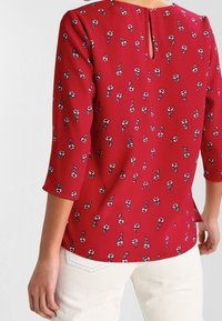 Blouse rouge avec un motif floral, manches trois-quarts et un détail dos en forme de goutte. Confectionnée dans un tissu léger à la texture lisse.