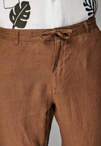 Pantalons en lin marron avec une coupe décontractée, dotés d'une taille à cordon, de poches latérales et d'une texture subtile. Sans motifs ni accents distincts.
