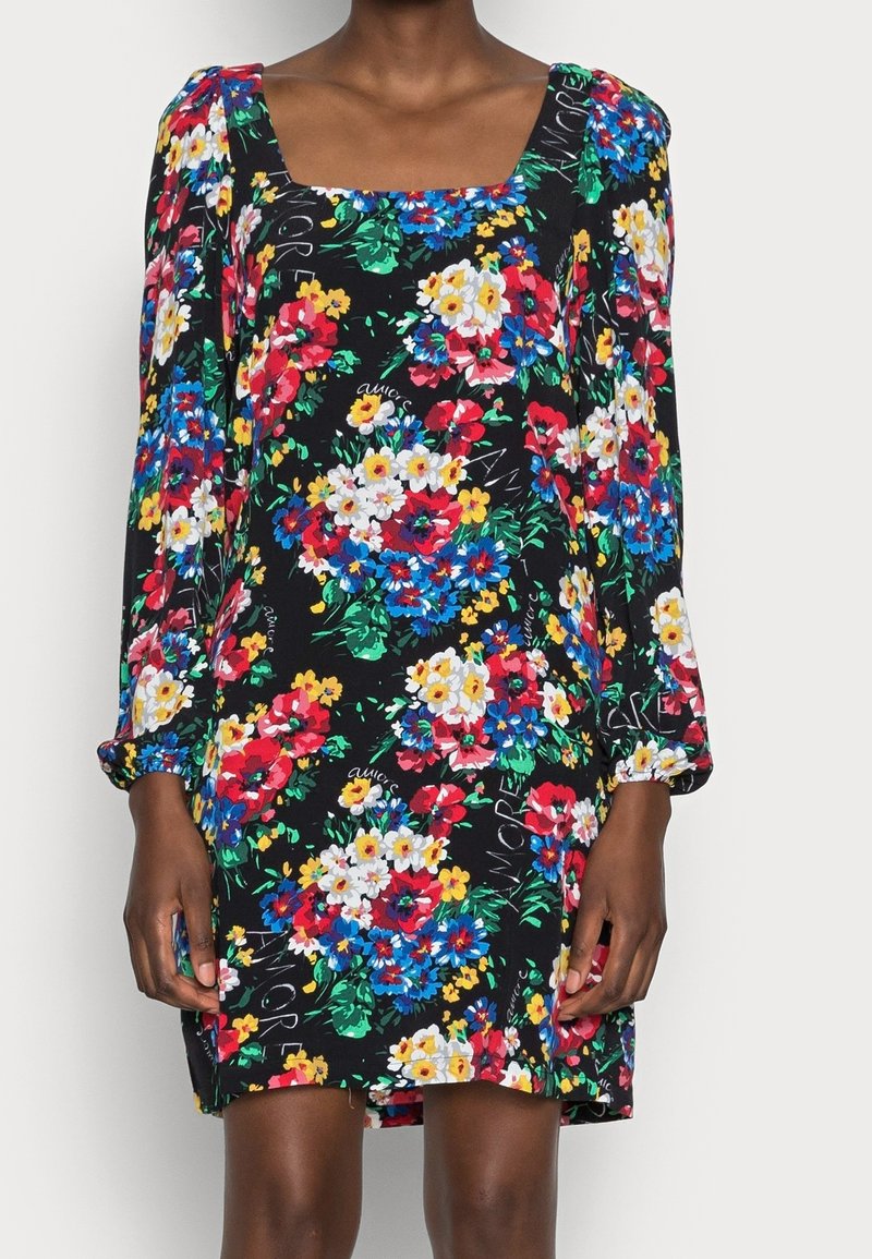 Vrouw in een zwarte jurk met lange mouwen, bedrukt met kleurrijke bloemen en met een vierkante halslijn, staande tegen een effen achtergrond.