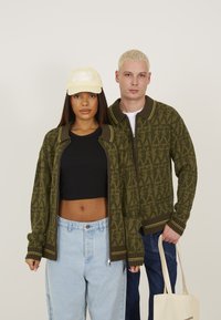 Olive green oversized knit cardigan with patterned design, ribbed cuffs, and contrasting yellow stripes; paired with light denim and a cap.