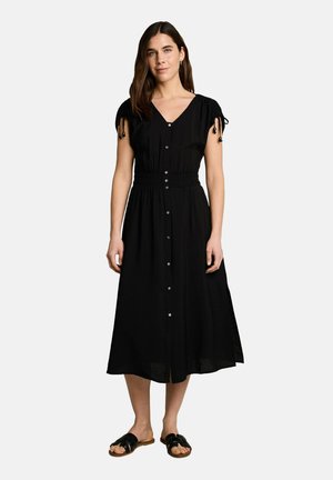 Frau steht und trägt ein schwarzes, knöpfbares Midikleid mit kurzen Ärmeln, die an den Schultern gebunden sind, und schwarze Slide-Sandalen vor einem schlichten Hintergrund.