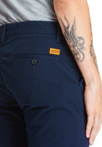 Mörkblå byxor i bomull, med en bakficka med knapp och en orange Timberland-logotyp. Synlig tatuering på armen.