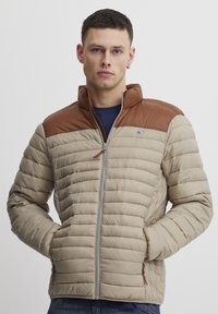 Beige pufferjakke med en brun skulderstykke, quiltet design, fuld lynlås og sidelommer; tekstureret stof med en glat finish.