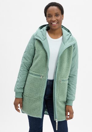 Lächende Person, die eine hellgrüne Fleecejacke mit Kapuze und gesteppte Ärmel über einem weißen Hemd und dunklen Jeans trägt.