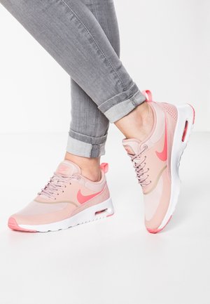 Fødder i opkørte grå jeans iført lyserøde og koralfarvede sporty sneakers med hvide såler på hvid baggrund.