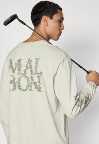 Long-sleeve beige shirt featuring green plant-themed text on the back and sleeve. Soft fabric with a relaxed fit. Golf club held behind the neck.