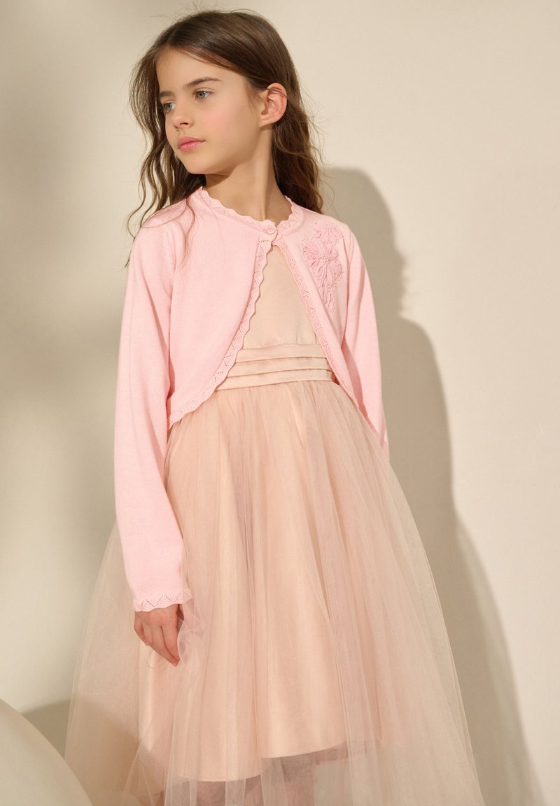 Cardigan en tricot rose avec bords festonnés, fermeture à boutons, porté sur une robe en tulle rose clair avec une jupe plissée, en tissu doux et léger.