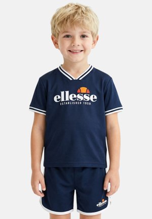 Garçon blond souriant, portant une tenue de sport ellesse bleu marine avec des bordures blanches et un logo sur le t-shirt et le short, debout sur un fond uni.