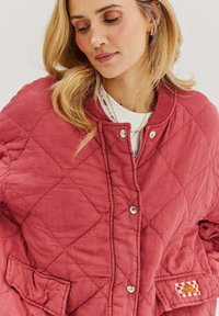 Prešívaná, hrdzavočervená bunda so striebornými cvočkami, bočnými vreckami a nášivkou s károvaným vzorom. Mäkčená, textúrovaná tkanina.