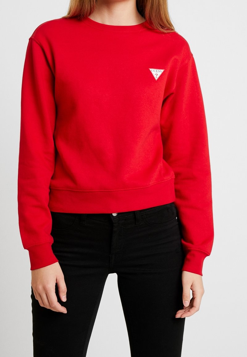 Personne portant un sweat-shirt rouge à manches longues avec un petit logo triangulaire blanc Guess et un pantalon noir sur un fond uni.