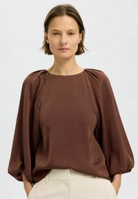 Blusa marrone con scollo rotondo e maniche a sbuffo. Il tessuto appare liscio e morbido, cadendo delicatamente sul torso.