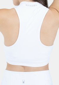 Biały sportowy crop top z designem racerback, wykonany z gładkiego materiału. Posiada małą metkę z logo przy kołnierzu i otwarty plecy.