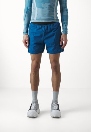 Blauwe sportshorts met een elastiche tailleband. Draagt een lichtblauw langemouwen shirt, grijze sokken, en unieke witte en grijze sneakers.