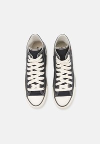 Čierne vysoké tenisky z plátna s bielymi gumovými podrážkami a šnúrkami. Majú biele šitie a bočné ventilačné otvory.