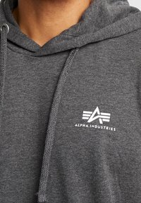 Szara bluza z kapturem wykonana z miękkiego materiału, z kieszenią typu kangur i kapturkiem ze sznurkiem. Biały logotyp na piersi z geometrycznym wzorem.