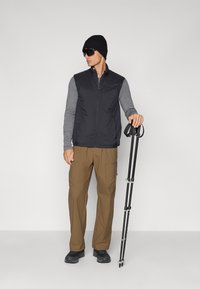 Un gilet isolant noir par-dessus une chemise à manches longues grise, un pantalon cargo marron, un bonnet noir, des lunettes de soleil sombres et tenant un bâton de trekking pliant.