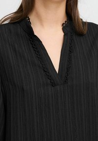 Haut noir avec un motif rayé texturé et un col en V. Présente une bordure décorative festonnée au niveau du col. Tissu doux.