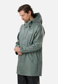 Uomo che sistema il polsino della manica di un impermeabile verde con cappuccio e tasche con bottoni, in piedi davanti a uno sfondo uniforme.