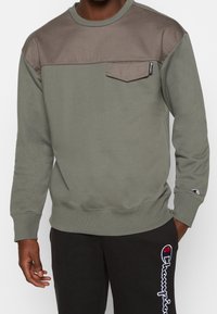 Uomo che indossa una felpa a maniche lunghe bicolore verde oliva e marrone con una tasca sul petto e pantaloni neri con testo del logo bianco e rosso.