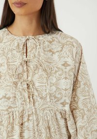 Beige blouse met een patroon van ingewikkelde draaiende ontwerpen. Beschikt over een ronde halslijn, gedeeltelijke knoopsluiting en eenGathered taille voor een vloeiende silhout.