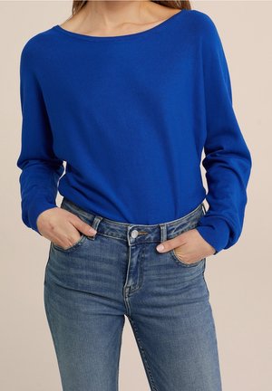 Femme portant un pull à manches longues bleu vif rentré dans un jean en denim bleu taille haute avec les mains dans les poches avant.