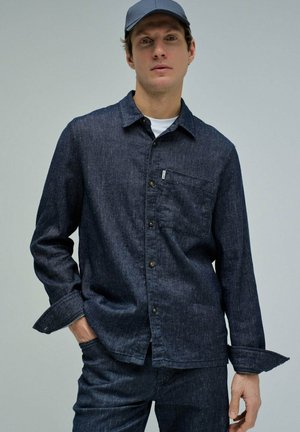 Hombre con camisa vaquera azul oscuro con bolsillo delantero y pantalones vaqueros a juego, gorra azul, de pie con una mano en el bolsillo.