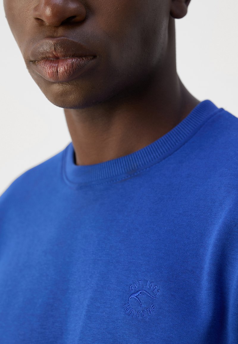 Royal blue sweatshirt wykonany z miękkiego materiału, z okrągłym żebrowanym dekoltem i małym haftowanym logo na piersi.