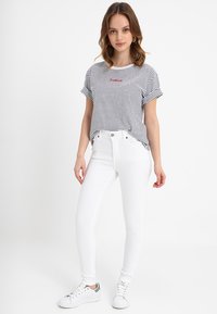 T-shirt de algodão às riscas em azul marinho e branco, com mangas curtas e um logótipo bordado em vermelho; combinado com jeans brancos ajustados e sapatilhas brancas.