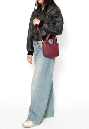 Vrouw die een zwarte leren jas en lichtblauwe wijde jeans draagt, met een kleine bordeauxrode leren handtas met schouderriem.