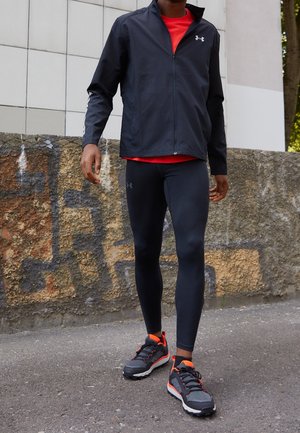 Uomo che cammina all'aperto indossando una giacca sportiva nera, leggings neri, maglietta rossa e scarpe da corsa nere con dettagli arancioni.